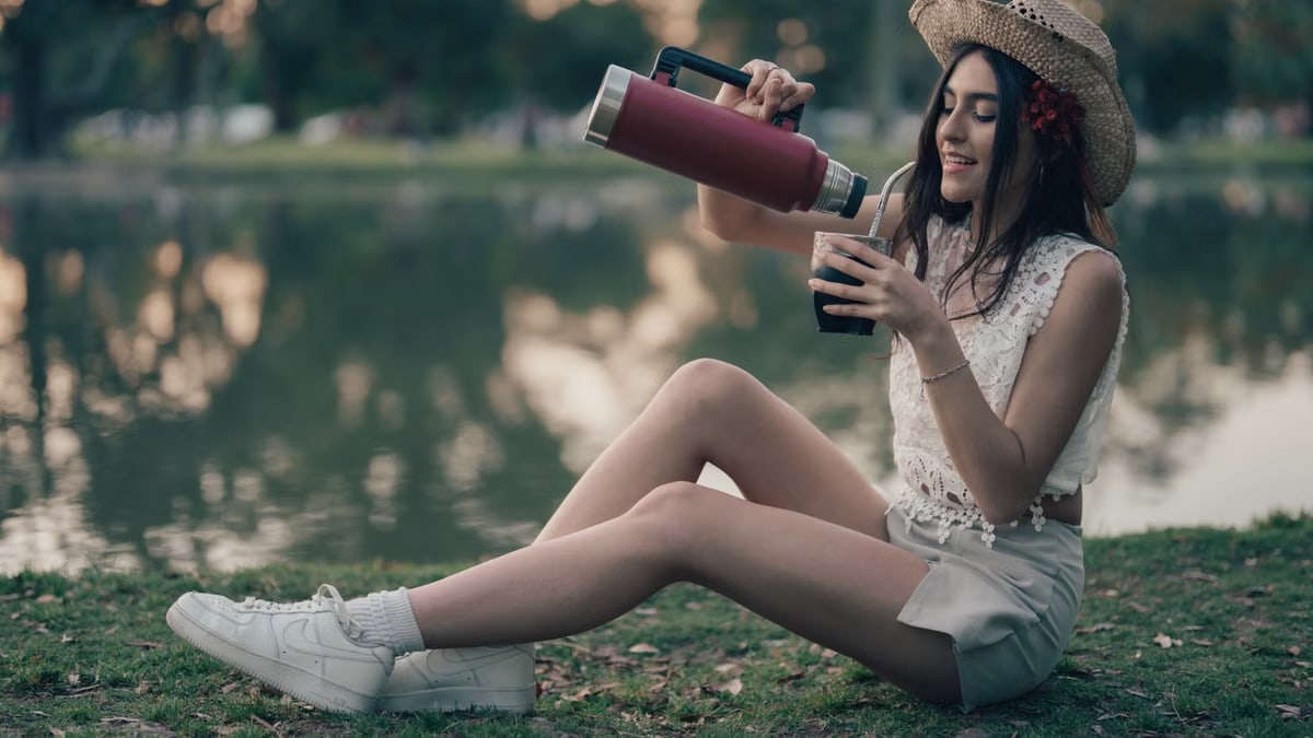 A young woman pours mate from a thermos, relaxing by a lake in Buenos Aires, Argentina.